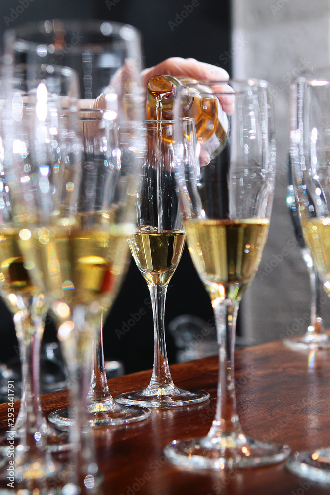 A large number of glasses with white wine at the bar. Wine is pouring into a glass. Wine tasting concept in a restaurant. Out of focus. Photo in the interior. A large number of glasses with white wine at the bar. Wine is pouring into a glass. Wine tasting concept in a restaurant. Out of focus. Photo in the interior.
