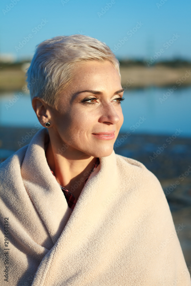 Adult middle-aged blonde woman wrapped herself in a bright plaid on the beach and looks at the sea, selective focus