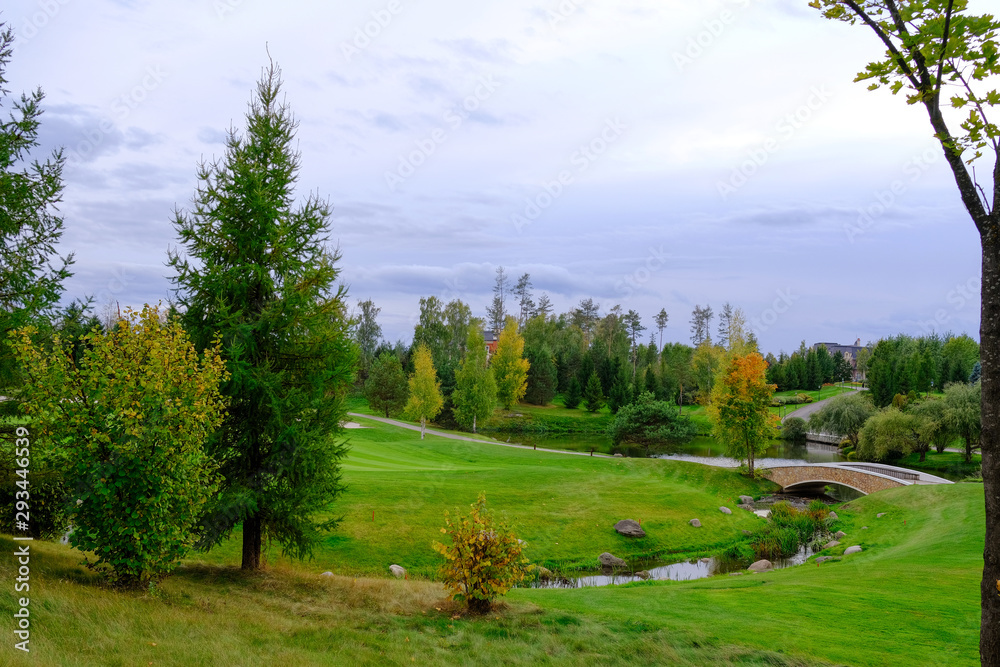 Autumn landscape. Hills with green lawn and ornamental shrubs and trees with a bridge over the creek against the evening sky. It's a beautiful park. Autumn landscape. Hills with green lawn and ornamental shrubs and trees with a bridge over the creek against the evening sky. It's a beautiful park.