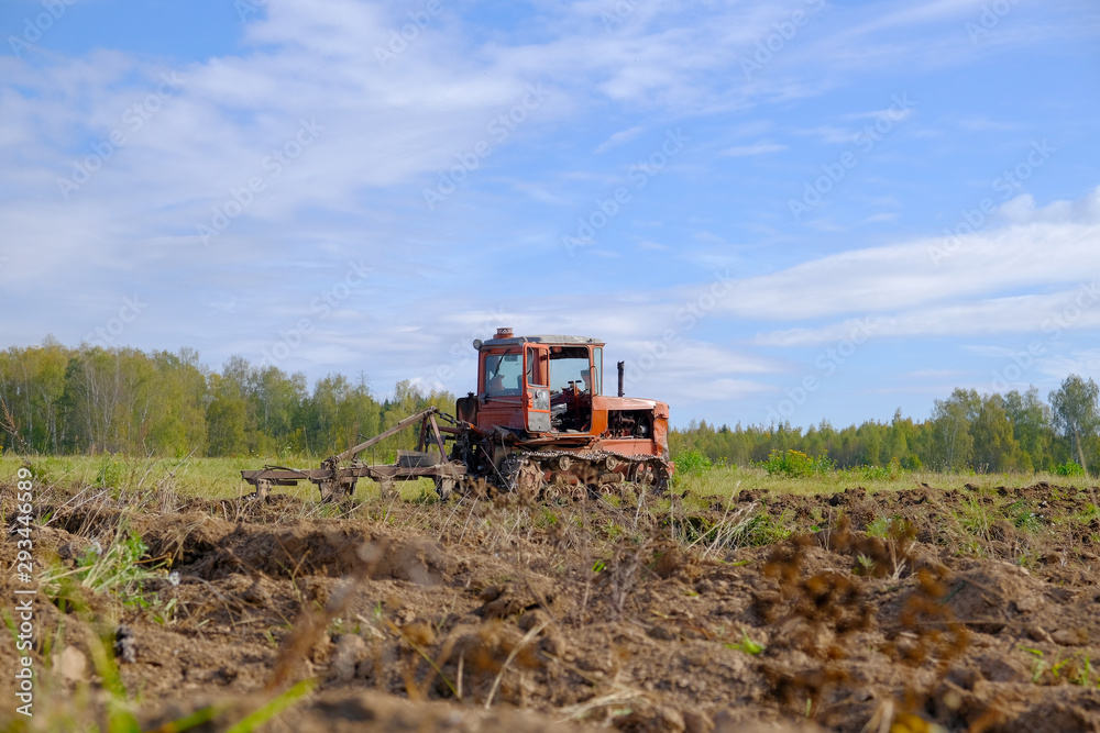 A tracked tractor with a plough plows the ground in autumn against the background of the forest.