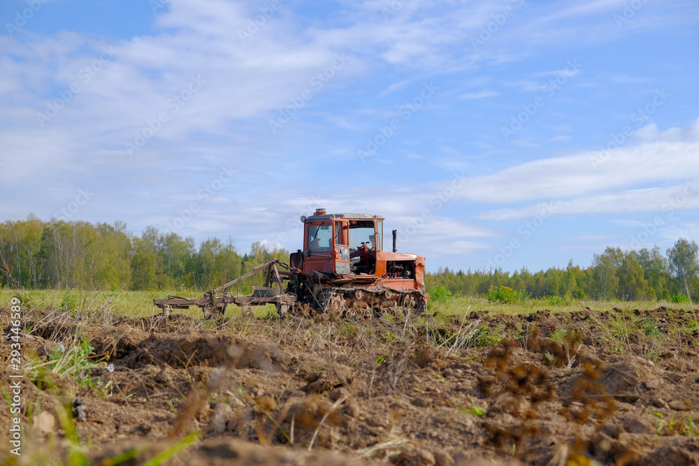 A tracked tractor with a plough plows the ground in autumn against the background of the forest. A tracked tractor with a plough plows the ground in autumn against the background of the forest.