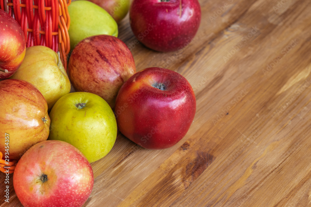 Red, green and yellow apples rolled out of a red wicker basket onto a brown wooden surface, side view from above Red, green and yellow apples rolled out of a red wicker basket onto a brown wooden surface, side view from above