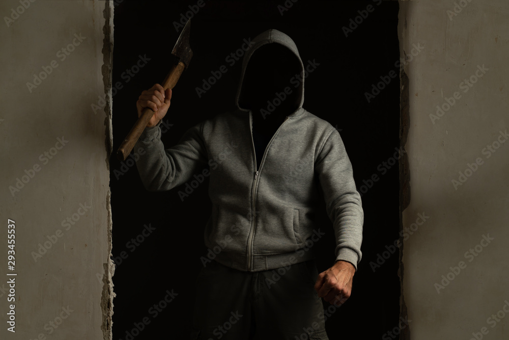A man with a hood on his head, swinging an axe, coming out of a dark room. Criminal with an axe