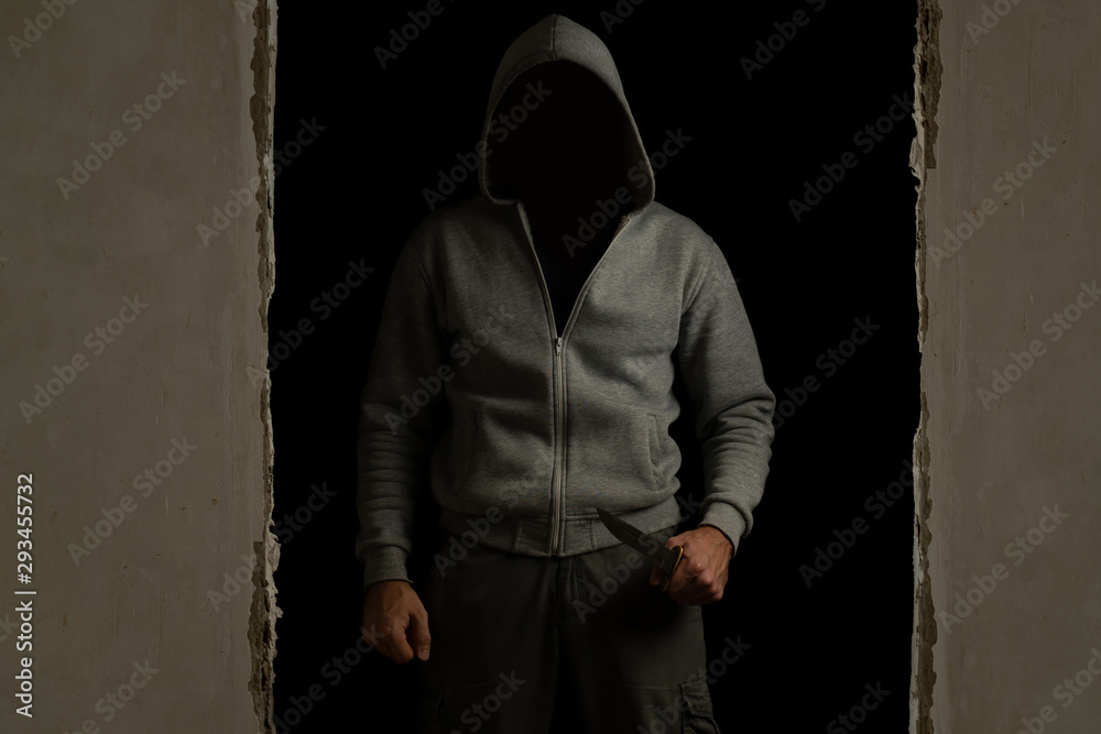 Unknown man with a hood on his head and a knife in his hand out of the dark room
