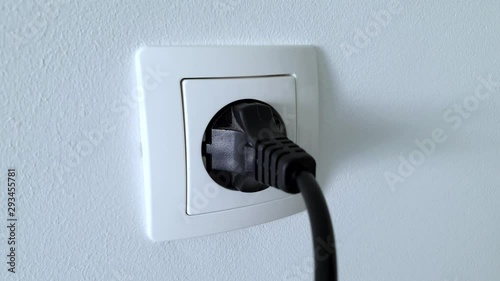 Connecting black electrical power plug with a hand inside a white plastic power socket on a white wall