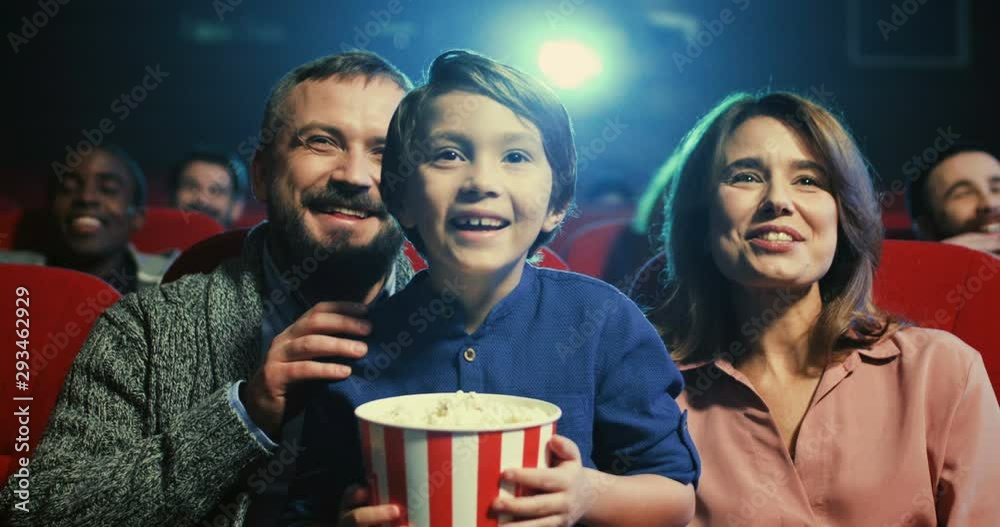 Portrait of teh happy small cute boy with popcorn in hands sitting in the cinema between cheerful parents and they laughing as watching comedy movie.
