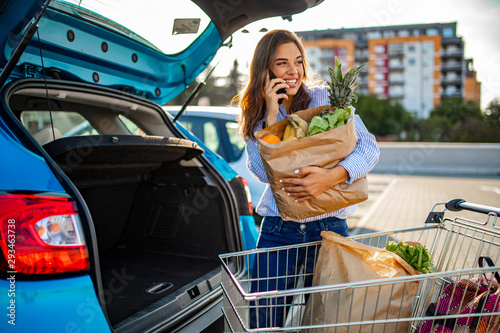 Foto Beautiful young woman shopping in a grocery store/supermarket, putting the groceries into her car in the parking lot, looking around and talking with smartphone