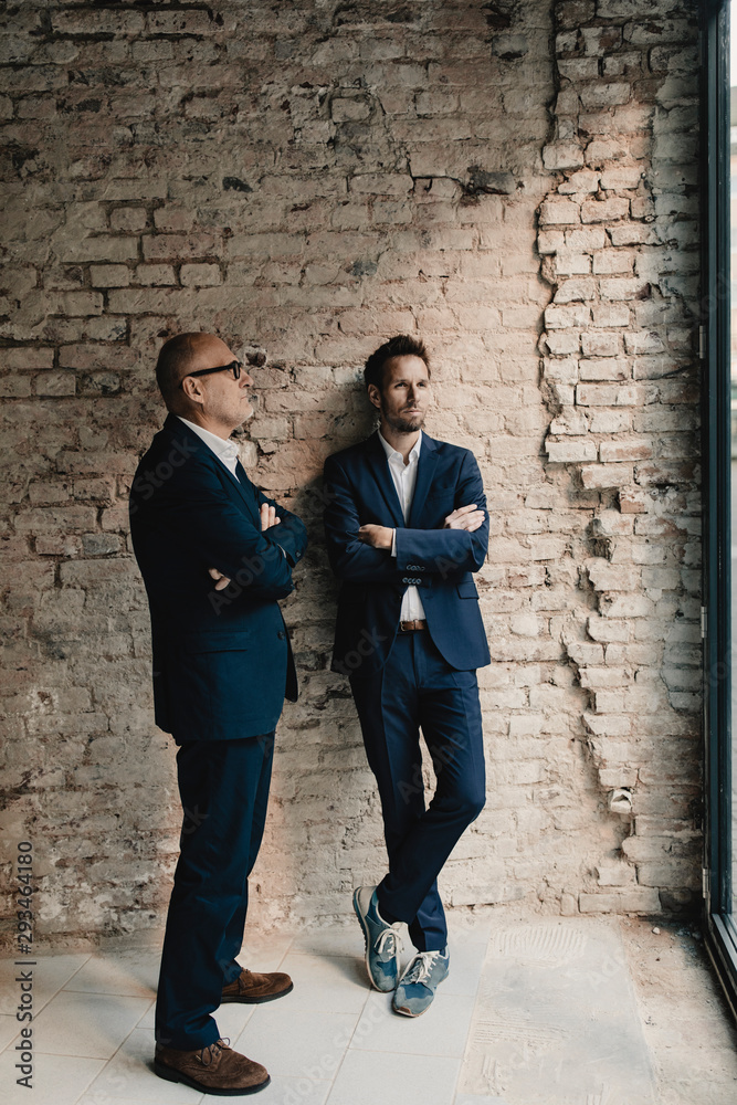Senior and mid-adult businessman standing at brick wall Senior and mid-adult businessman standing at brick wall