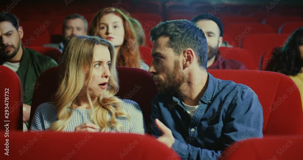 Caucasian young attractive boyfriend and girlfriend sitting together in the cinema and fighting because of the popcorn and people from behind rows asking them to be quietly. Caucasian young attractive boyfriend and girlfriend sitting together in the cinema and fighting because of the popcorn and people from behind rows asking them to be quietly.