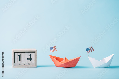 wooden calendar with October 14 date near paper boats with American flag pattern on blue background