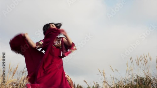 Gipsy woman in beautiful red dress is dancing with smile in field. Blue sky on the background