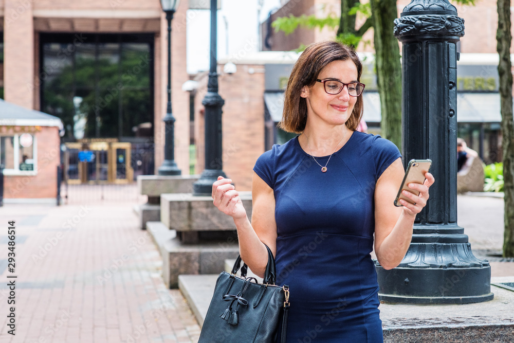 Young 40 years old Native American Teacher working in New York, wearing short sleeve dress, glasses, arm carrying bag, standing on street by light poles on school campus, reading messages on phone..