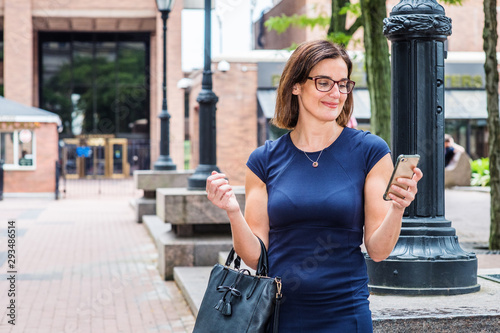 Obraz na płótnie Young 40 years old Native American Teacher working in New York, wearing short sleeve dress, glasses, arm carrying bag, standing on street by light poles on school campus, reading messages on phone.. w salonie