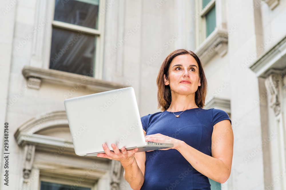 Young 40 years old Native American Businesswoman working in New York City, wearing short sleeve dress, holding laptop computer, standing outside office building with windows, looking up, thinking.. Young 40 years old Native American Businesswoman working in New York City, wearing short sleeve dress, holding laptop computer, standing outside office building with windows, looking up, thinking..