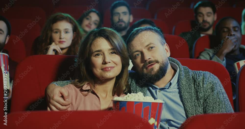 Portrait shot of the Caucasian good looking married couple, man and woman sitting together in the cinema with popcorn, hugging and watching movie. Portrait shot of the Caucasian good looking married couple, man and woman sitting together in the cinema with popcorn, hugging and watching movie.