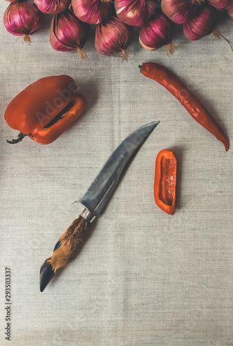 hunting knife with a handle in the form of a goat's hoof with red bell pepper, chili pepper and a bunch of onions on the background of a linen tablecloth