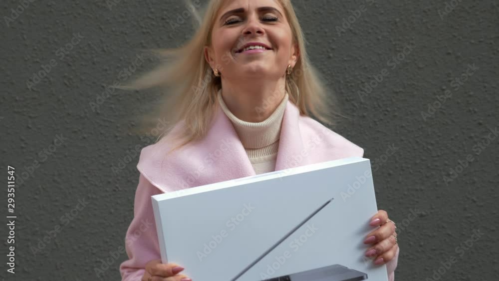 Young attractive blonde woman in pink coat is shopping. The lady holds laptop package in her hands, smiles and jumps from happiness. Christmas holidays, sale retail industry concept. Close-up. 4K Young attractive blonde woman in pink coat is shopping. The lady holds laptop package in her hands, smiles and jumps from happiness. Christmas holidays, sale retail industry concept. Close-up. 4K