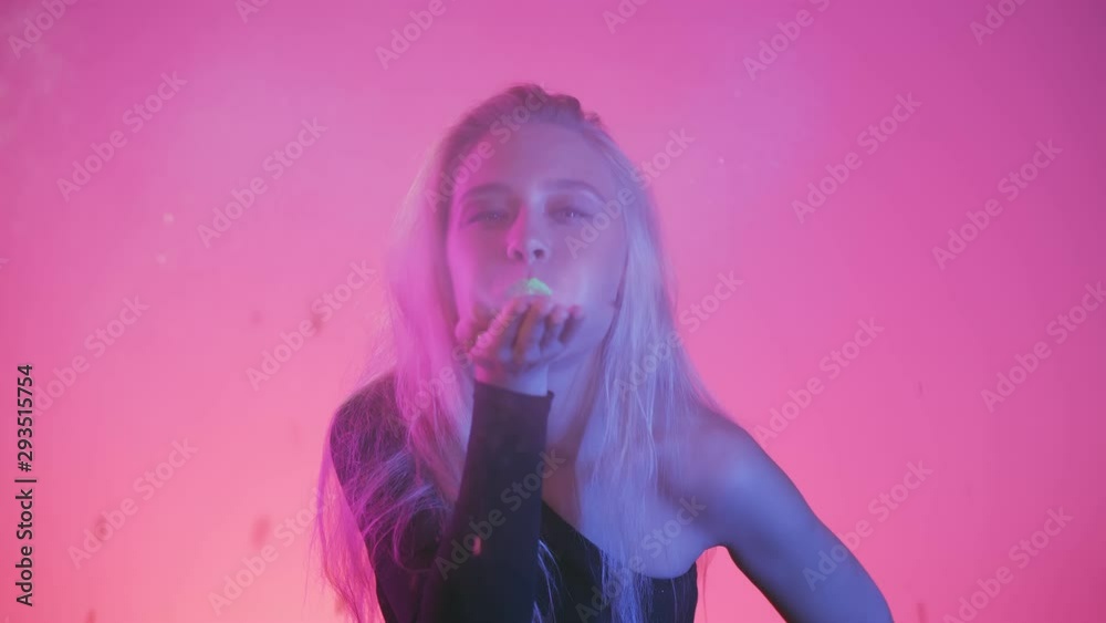 Vivid neon style light. Sexy young woman with with creative make up art face blowing confetti glitter or pieces of gold foil on the camera, it shines brightly in the light