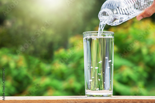 Hand pouring drinking water from bottle into glass on blurred green nature background