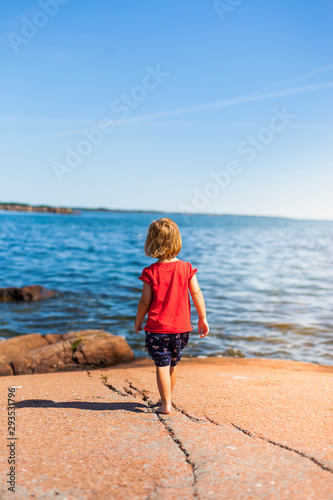 Kind bewundert Meerblick. Kleines Kind geht auf rotem Felsen am Meeresufer. Kid standing on red rock looking on the ocean. Scandinavian sea view.