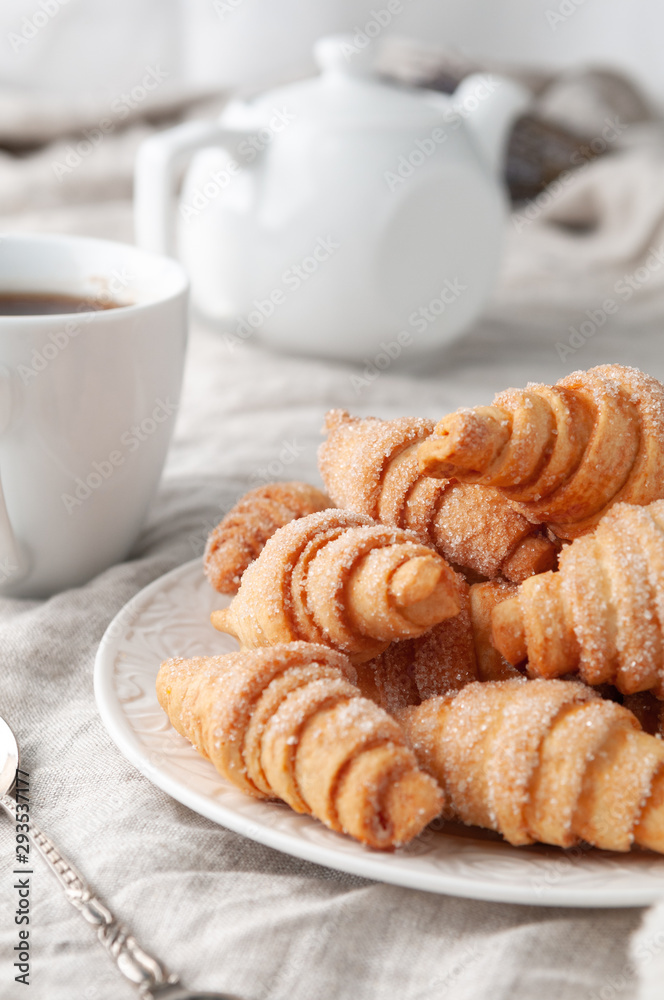 Homemade roll-shaped shortbread. Sprinkled with sugar. On a white plate. Background gray linen. In the background is a white teapot and a cup. Country style. Homemade roll-shaped shortbread. Sprinkled with sugar. On a white plate. Background gray linen. In the background is a white teapot and a cup. Country style.