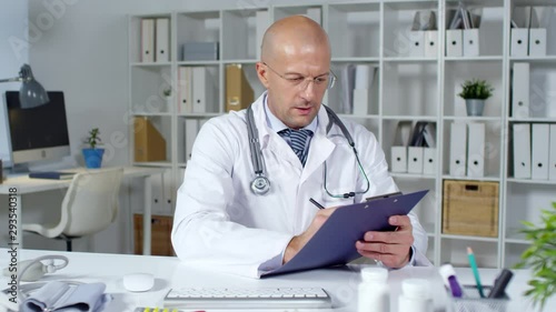 Wallpaper Mural Medium shot of bald male physician in white coat sitting at his desk and writing notes on clipboard while consulting patient via video call Torontodigital.ca