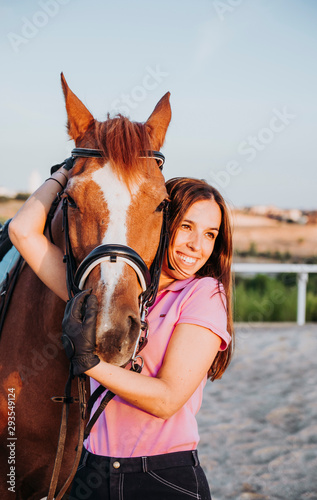 Young smiling woman standing in relax outside with wonderful chestnut horse on bridle and looking away.