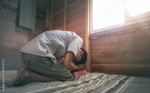 Young male kneel down and praying on holy Bible with morning light in bed room. christian concept. © surachat