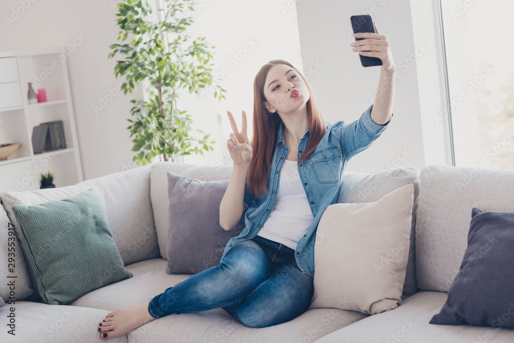 Photo of flirty lady telephone in hands making selfies showing v-sign symbol sending air kisses sitting comfy on sofa wearing jeans clothes apartment indoors Photo of flirty lady telephone in hands making selfies showing v-sign symbol sending air kisses sitting comfy on sofa wearing jeans clothes apartment indoors