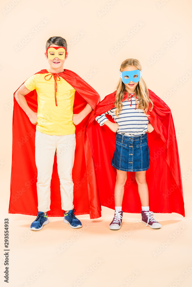 full length view of two smiling kids in masks and red cloaks standing with hands on hips on pink