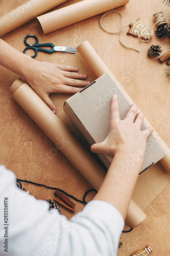 Wrapping christmas gifts concept. Hands wrapping christmas gift in craft paper and pine branches, cones, gingerbread cookies, thread, cinnamon, scissors on rural wooden table