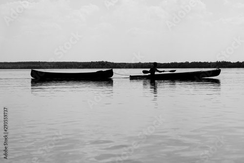 Mann rudert mit einem Boot über großen See und schleppt ein Kanu ab, in Schwarz Weiß.