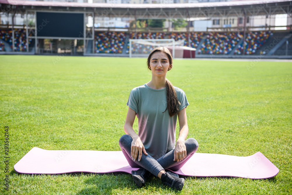 Fototapeta premium Woman training outdoors. She's sitting on mat on green grass and doing yoga exercises