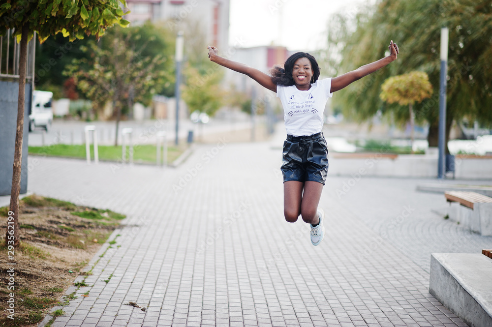 African american dark skinned slim model posed in a black leather shorts and white t-shirt. She jumping at the air.