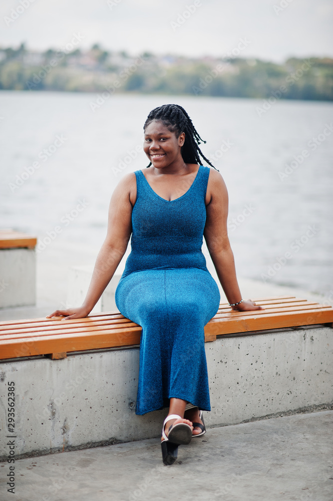African american dark skinned plus size model posed in a blue shiny dress against sea side sitting on bench. Stock Adobe Stock