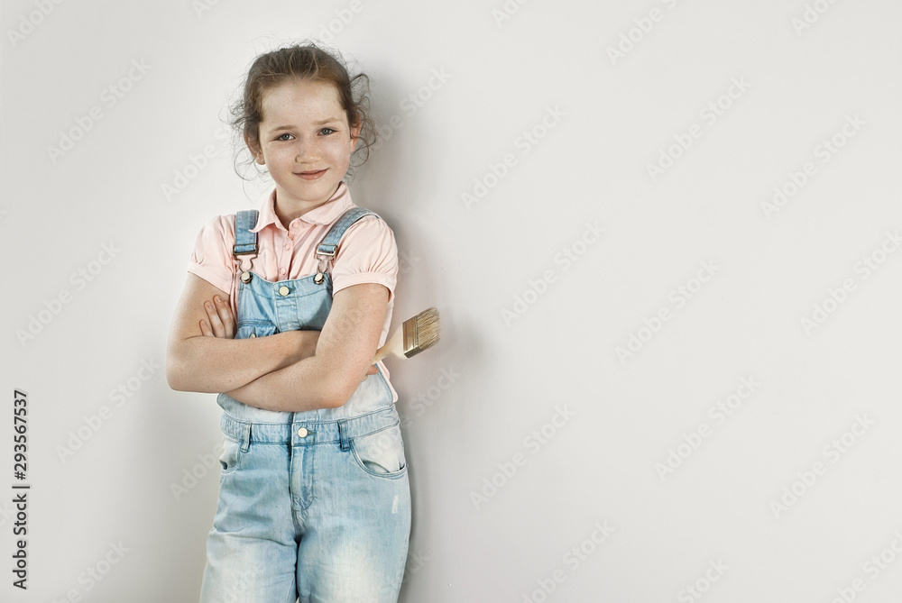 Little girl makes repairs in blue overalls. The child paints the walls. Girl with brush in hands on white wall background. Little girl makes repairs in blue overalls. The child paints the walls. Girl with brush in hands on white wall background.