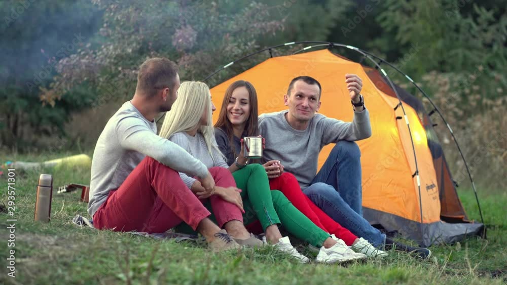 Four friends in the forest are sitting and talking on the grass near the tent and campfire. The guy emotionally tells the story, waving his arms. smoke from the fire flies past. Four friends in the forest are sitting and talking on the grass near the tent and campfire. The guy emotionally tells the story, waving his arms. smoke from the fire flies past.
