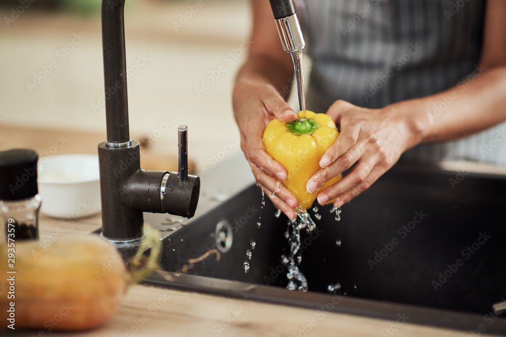 Close up of mixed race woman in apron standing in kitchen ad washing yellow paprika in sink. - obrazy, fototapety, plakaty 