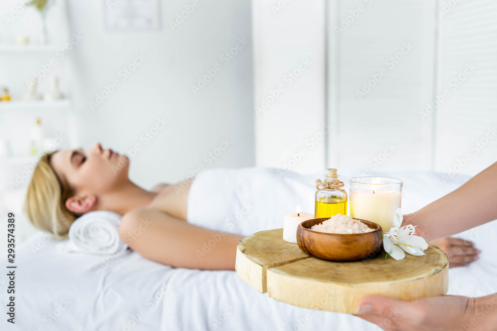 selective focus of masseur holding board with fragrance oil, candles, sea salt and flower