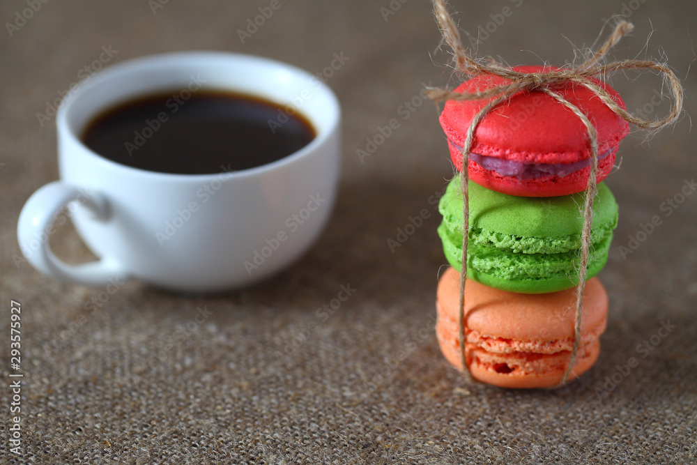 Macaron three pieces red, green and orange tied with rope and a mug of coffee on burlap tablecloth. Macaron three pieces red, green and orange tied with rope and a mug of coffee on burlap tablecloth.