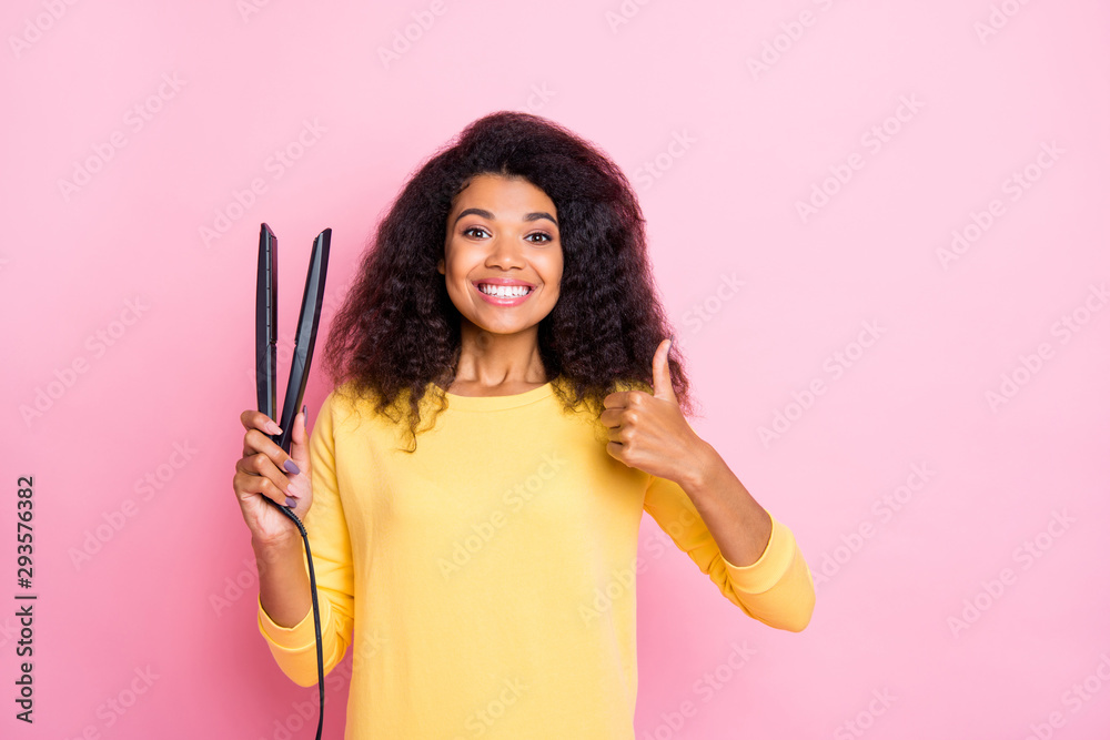 Portrait of positive cheerful black girl hold hair straightener enjoy her new professional treatment show thumb-up sign wear yellow jumper isolated over pink color background