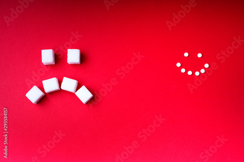  The choice of sweetener in tablets or regular sugar. An alternative to sugar for people with diabetes. On a red background, cubes of white sugar with a sad smiley and sugar substitute cheerful smiley