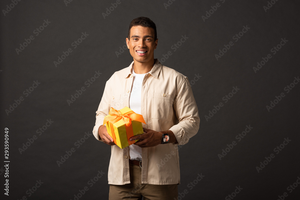 smiling handsome mixed race man in beige shirt holding present on black background