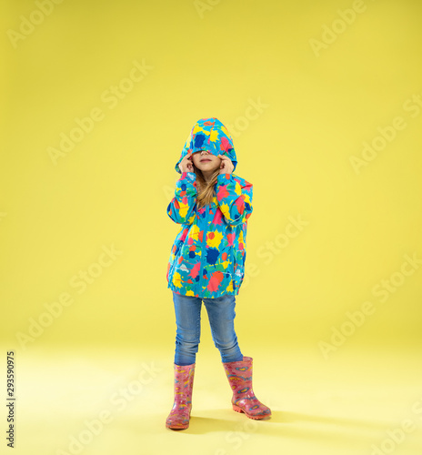 A full length portrait of a bright fashionable girl in a raincoat holding an umbrella of rainbow colors on yellow studio background. Autumn and spring fashion for kids. Cute stylish blonde girl.