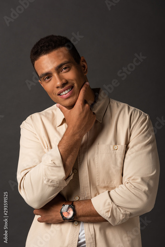 smiling handsome mixed race man in beige shirt touching face on black background