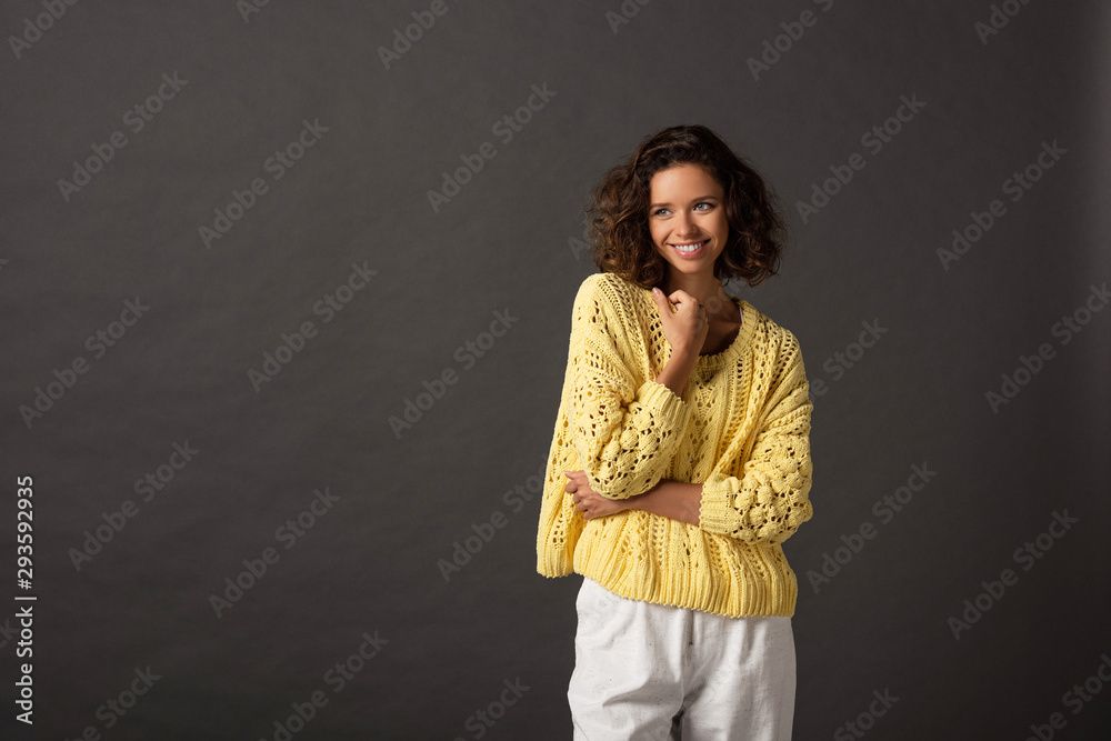 smiling curly woman in yellow knitted sweater looking away on black background