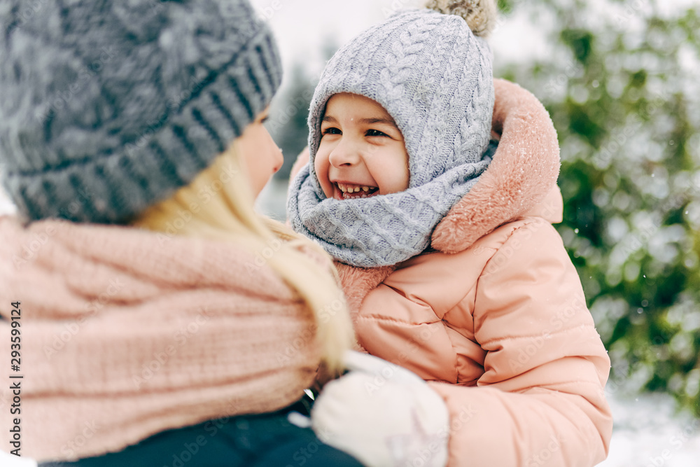 Rear view close-up portrait of happy little girl smiling and playing together with her mother in the park on snowing day. Beautiful woman and her child walking on winter day. Christmas mood. Childhood Rear view close-up portrait of happy little girl smiling and playing together with her mother in the park on snowing day. Beautiful woman and her child walking on winter day. Christmas mood. Childhood