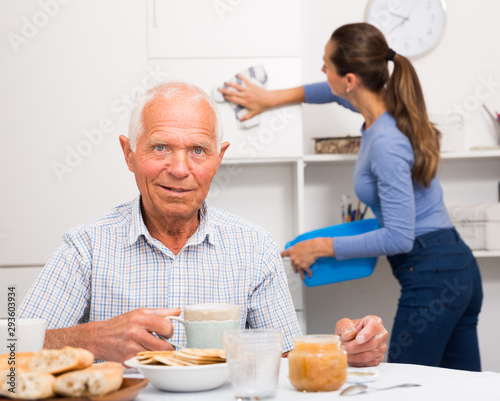 Elderly man drinkimg tea, daughter washing furniture kitchen at home
