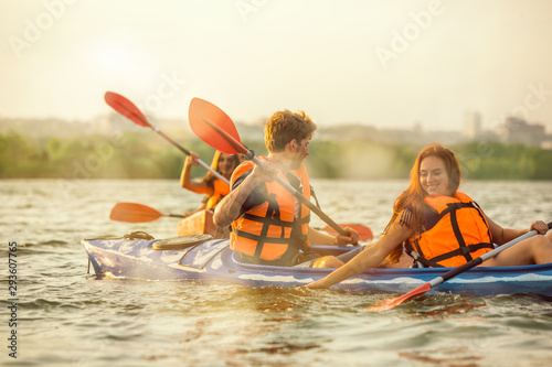 Happy young caucasian group of friends kayaking on river with sunset in the backgrounds. Having fun in leisure activity. Happy male and female model laughting on the kayak. Sport, relations concept.
