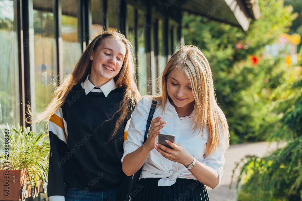 Fototapeta premium Happy moments of two stylish girls hugging and looking at the phone, discussing something on the street in the city. Close up portrait of cheerful joyful attarctive young girls best friends.